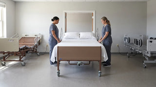 Two care staff preparing an expandable bariatric hospital bed with clear working access on both sides in a long-term care room