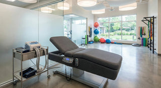 Interior of a modern physical therapy clinic featuring a sleek gray lumbar traction table and a medical cart with a cold compression device in the foreground, with a bright rehabilitation gym area containing stability balls in the background.