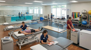 A spacious physical therapy clinic featuring treatment tables, rehabilitation equipment, and a hydrotherapy pool, with clinicians assisting patients across different therapy stations.