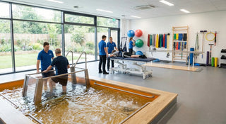 A bright rehabilitation clinic with a hydrotherapy pool in use, therapists assisting patients on treatment tables, and various rehab tools such as resistance bands, stability balls, and parallel bars arranged along the wall.