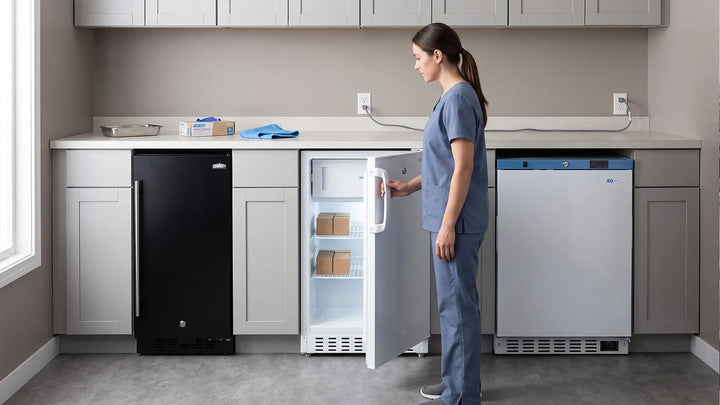 Practice nurse at an open undercounter refrigerator in a clinical cabinet run showing three built-in units of increasing width