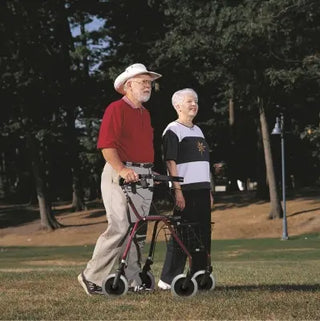 Two elderly individuals walking outdoors with a Clarke Healthcare D12050X Dolomite Legacy Standard Walker on a grassy area.