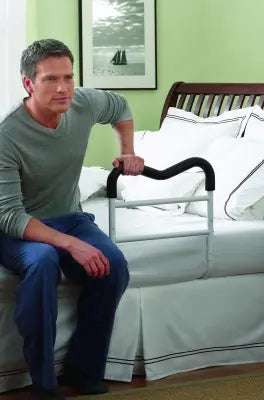 Man sitting on a bed holding a black white Clarke Healthcare HMR400NW M-Rail Bed Rail handle, with a framed picture on the wall.