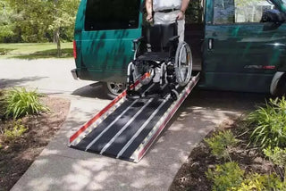 Person using a wheelchair with a Clarke Healthcare MPEBXX DECPAC Fiberglass Portable Wheelchair Van Ramp with Edge Barrier attached to a green van in a park-like setting.