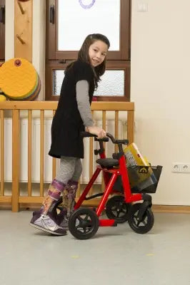 Child pushing a Clarke Healthcare S760XXXX Marcy Telescoping Rollator for Pediatrics indoors with a wooden fence and wall in the background