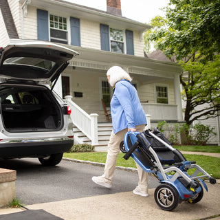 An older woman carrying a folded Journey So Lite 08474 scooter across a driveway towards an open car trunk.