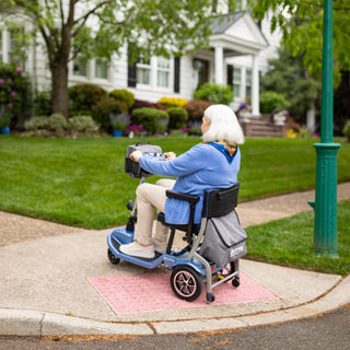 A woman with white hair riding a Journey So Lite 08474 scooter along a pavement past a white house.