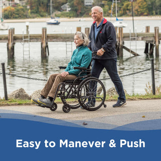 A man pushing a woman seated in a Journey So Lite C2 08970 wheelchair along a waterfront with boats behind them.