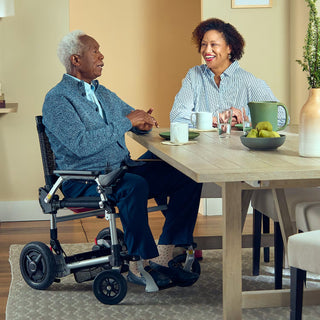 A man seated in a Journey Zoomer 09017 power chair at a dining table, talking with a woman over coffee.