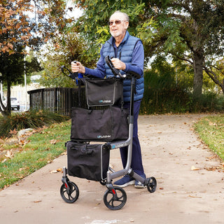 An older man in sunglasses and a blue vest walking on a paved path with the Journey UPWalker EZ Lite and its storage bags.