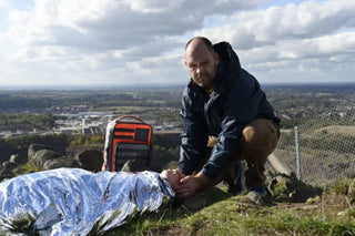 Person tending to a person wrapped in a mylar blanket on a hillside with a scenic view.
