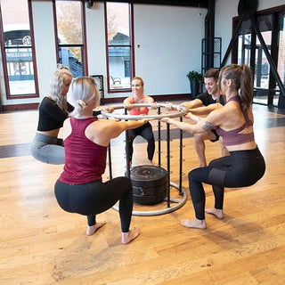 The Stroops CONNECTUS is a black and gray circular steel fitness training system with a round barre at the top and six vertical anchor spines arranged around the base. Each spine provides multiple connection points for resistance bands, allowing up to six people to perform full-body training around the system.