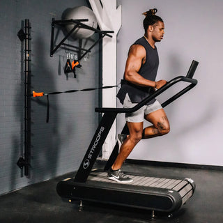 A black flat self-propelled motorless treadmill with a low-profile steel frame, slatted running belt, front support handles, and a vertical steel spine positioned behind the treadmill. The system can be configured with optional resistance training accessories including the Base Slastix® Adjustable Anchor, Universal Swivel Belt XL, and Optitail® Anchoring System for performance training and rehabilitation applications.
