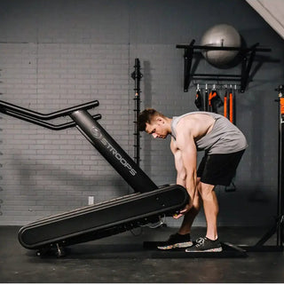 A black flat self-propelled motorless treadmill with a low-profile steel frame, slatted running belt, front support handles, and a vertical steel spine positioned behind the treadmill. The system can be configured with optional resistance training accessories including the Base Slastix® Adjustable Anchor, Universal Swivel Belt XL, and Optitail® Anchoring System for performance training and rehabilitation applications.
