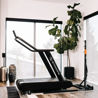 A black flat self-propelled motorless treadmill with a low-profile steel frame, slatted running belt, front support handles, and a vertical steel spine positioned behind the treadmill. The system can be configured with optional resistance training accessories including the Base Slastix® Adjustable Anchor, Universal Swivel Belt XL, and Optitail® Anchoring System for performance training and rehabilitation applications.