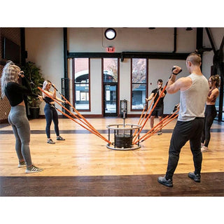 The Stroops REVOL Revolution Anchor is a circular black group fitness resistance system with six vertical pillars arranged around a central swiveling anchor structure. Multiple anchor points are positioned at different heights around the system for attaching resistance bands and supporting movement-based group training.