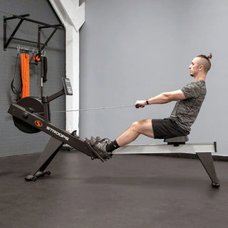 The Stroops SXR Xtreme Rower is a commercial rowing machine with a black flywheel housing, long aluminum monorail, ergonomic padded seat, adjustable footrests, and an angled padded handle. A user-friendly performance console is mounted above the flywheel, while integrated transport wheels and a foldable frame allow the rower to be moved and stored easily when not in use.