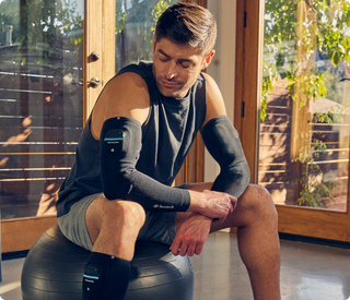 Man on an exercise ball wearing the Therabody RecoveryPulse Arm sleeve, with a RecoveryPulse Calf sleeve on his leg