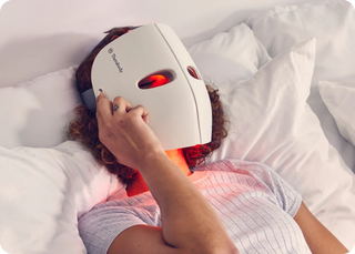 Person lying in bed wearing the Therabody TheraFace Mask with red light showing through the eye openings