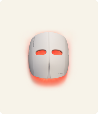 Front view of the Therabody TheraFace Mask glowing red against a cream background