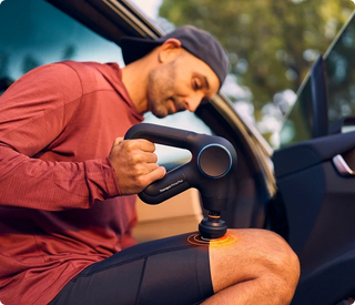 Man using the Therabody Theragun Prime Plus with the heated head on his thigh beside a car